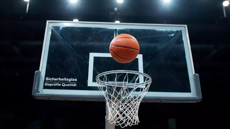Basketball im Flug vor dem Korbring mit weißem Netz und transparentem Plexiglas-Backboard
